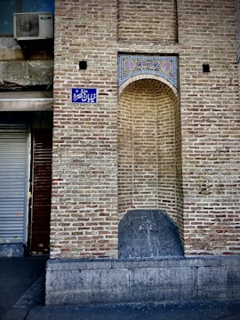 A brick wall featuring a decorative arch with intricate tile work above it, set in an urban environment. An air conditioning unit is mounted on the upper left, and a blue street sign with white lettering is affixed to the wall. The base of the wall has a stone bench-like structure integrated into it, displaying signs of weathering.