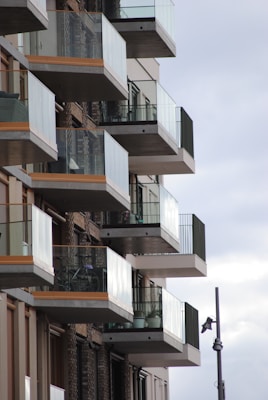 A series of modern apartment balconies with glass railings are aligned in a staggered fashion. The building has a contemporary design, featuring a combination of concrete and brick elements. A bicycle is visible on one of the balconies, and a streetlight is positioned nearby.