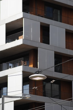 A modern apartment building with a focus on a streetlight suspended by cables. The building features a mix of white and wooden panels with balconies enclosed by glass railings.