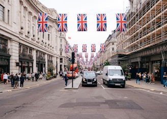 A busy urban street scene with a row of Union Jack flags hanging overhead. The street is lined with historic buildings, one under renovation with scaffolding. Pedestrians are walking along the sidewalks and vehicles are moving on the road, including a black taxi and a white van.