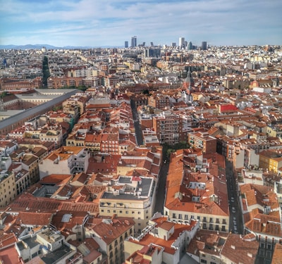 Aerial view of an urban area with densely packed buildings featuring red-tiled roofs. Streets separate different blocks of residential and commercial structures. The background showcases a city skyline with several tall skyscrapers under a partly cloudy sky.