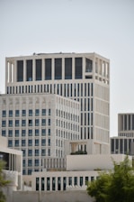 A cluster of modern high-rise buildings with a minimalist architectural style featuring vertical lines and large windows. The structures are primarily light-colored, with a clear sky as the background. Green foliage is visible in the lower foreground, suggesting an urban environment with some natural elements.