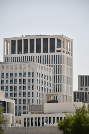 A cluster of modern high-rise buildings with a minimalist architectural style featuring vertical lines and large windows. The structures are primarily light-colored, with a clear sky as the background. Green foliage is visible in the lower foreground, suggesting an urban environment with some natural elements.