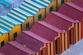 A row of beach huts with alternating yellow and red striped designs. The roofs are red, and a few objects, possibly small items like locks or toys, are placed on top of them. On the side, a deck area is visible with striped sun loungers matching the yellow and green color scheme of some huts.