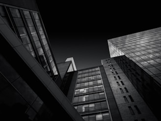 A dramatic black and white photograph depicting modern, high-rise buildings from a low-angle perspective. The image captures the geometric lines and reflective glass surfaces of the architecture, creating a sense of scale and depth against a dark sky.