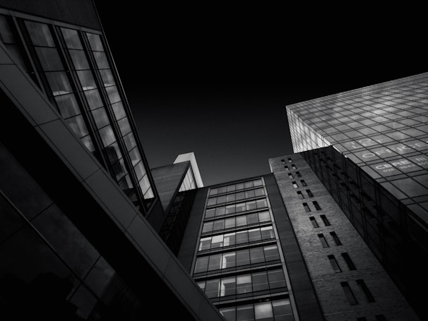 A dramatic black and white photograph depicting modern, high-rise buildings from a low-angle perspective. The image captures the geometric lines and reflective glass surfaces of the architecture, creating a sense of scale and depth against a dark sky.