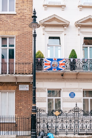 A building facade displays a mix of traditional brick and white plaster architecture. A large Union Jack flag decorates the front balcony, which also features metal railings. A street sign reads 'Charles Street W1 City of Westminster.' Below the balcony, there is an ornate black iron gate with gold detailing, and a lamppost stands adjacent. Green topiary plants are visible on the balcony. Blue plaques and additional buildings can be seen in the background.