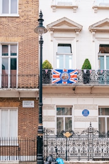 A building facade displays a mix of traditional brick and white plaster architecture. A large Union Jack flag decorates the front balcony, which also features metal railings. A street sign reads 'Charles Street W1 City of Westminster.' Below the balcony, there is an ornate black iron gate with gold detailing, and a lamppost stands adjacent. Green topiary plants are visible on the balcony. Blue plaques and additional buildings can be seen in the background.