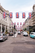 A busy urban street is decorated with numerous Union Jack flags hanging above. The street is lined with historic buildings on each side, showcasing classic architectural details. Cars, taxis, and a red double-decker bus are visible, along with pedestrians walking along the sidewalks. Scaffolding is present on one of the buildings, indicating construction or renovation work.