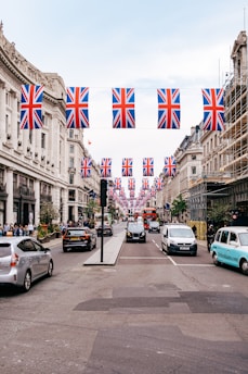 A busy urban street is decorated with numerous Union Jack flags hanging above. The street is lined with historic buildings on each side, showcasing classic architectural details. Cars, taxis, and a red double-decker bus are visible, along with pedestrians walking along the sidewalks. Scaffolding is present on one of the buildings, indicating construction or renovation work.