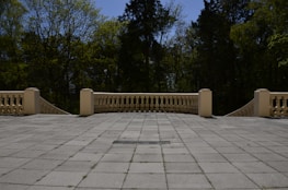 A spacious, tiled terrace foreground bordered by a light-colored balustrade, with a backdrop of lush, dense trees under a clear blue sky.