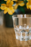 A clear glass tumbler is prominently placed on a smooth wooden surface. In the blurred background, there are vibrant yellow flowers, suggesting a soft and calm setting.
