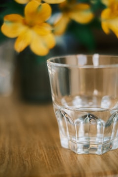 A clear glass tumbler is prominently placed on a smooth wooden surface. In the blurred background, there are vibrant yellow flowers, suggesting a soft and calm setting.