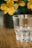 A clear glass tumbler is prominently placed on a smooth wooden surface. In the blurred background, there are vibrant yellow flowers, suggesting a soft and calm setting.