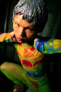 A person with a silver, glittery wig poses while wearing a vibrant, multicolored outfit featuring shades of red, yellow, green, and blue. The setting appears dim, with focused lighting emphasizing the person's intense gaze and striking appearance.