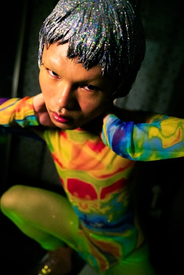 A person with a silver, glittery wig poses while wearing a vibrant, multicolored outfit featuring shades of red, yellow, green, and blue. The setting appears dim, with focused lighting emphasizing the person's intense gaze and striking appearance.
