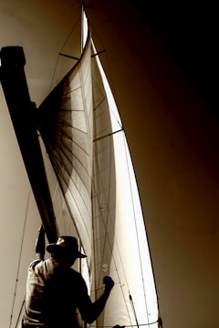 A person wearing a hat is silhouetted against a large sail. The light and shadow create a dramatic contrast on the sailcloth, highlighting its texture and structure. The angle suggests the person is involved in handling or observing the sail, contributing to a nautical theme.
