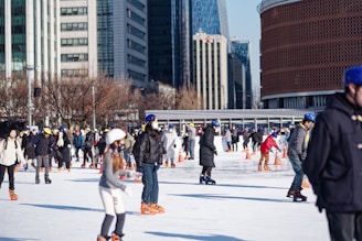 A bustling ice skating rink in an urban area with numerous people of various ages skating. Many skaters are wearing helmets and winter clothing. Surrounding the rink are tall buildings and bare trees, suggesting a city setting in colder weather.