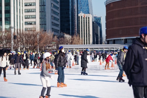 A bustling ice skating rink in an urban area with numerous people of various ages skating. Many skaters are wearing helmets and winter clothing. Surrounding the rink are tall buildings and bare trees, suggesting a city setting in colder weather.