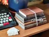A collection of vintage books stacked on a wooden table, tied together with twine. An old typewriter with black and white keys is nearby, and a few pieces of paper or tickets are scattered beside the books. A blue pot containing a plant with orange flowers is partially visible.