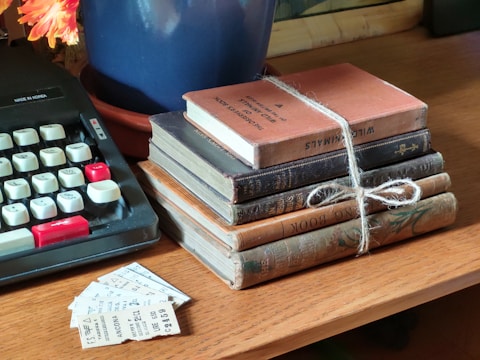 A collection of vintage books stacked on a wooden table, tied together with twine. An old typewriter with black and white keys is nearby, and a few pieces of paper or tickets are scattered beside the books. A blue pot containing a plant with orange flowers is partially visible.