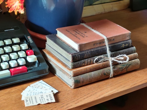 A collection of vintage books stacked on a wooden table, tied together with twine. An old typewriter with black and white keys is nearby, and a few pieces of paper or tickets are scattered beside the books. A blue pot containing a plant with orange flowers is partially visible.