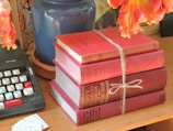 A stack of red hardcover books tied together with a string, placed on a wooden surface. Adjacent to the books, there is a vintage typewriter with white and red keys. A blue ceramic pot and vibrant orange flowers can be seen in the background, adding a decorative touch to the scene.