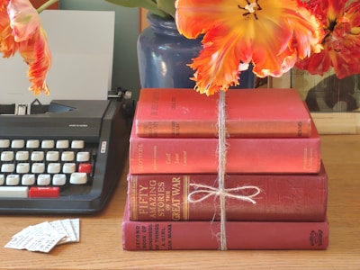 A stack of four red books tied with twine rests on a wooden surface. To the left, there is a vintage typewriter with white and red keys, and several small paper cuttings are placed nearby. Above the books, vivid orange and yellow flowers, likely tulips, emerge from a blue vase.