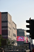 A modern urban scene with geometrically patterned buildings under a clear sky. The structures have a mix of red and gray tones with distinctive diamond shapes. A large digital screen displays vibrant red and white content. The foreground includes silhouetted trees, traffic lights, and people moving along the street.