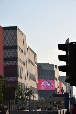 A modern urban scene with geometrically patterned buildings under a clear sky. The structures have a mix of red and gray tones with distinctive diamond shapes. A large digital screen displays vibrant red and white content. The foreground includes silhouetted trees, traffic lights, and people moving along the street.