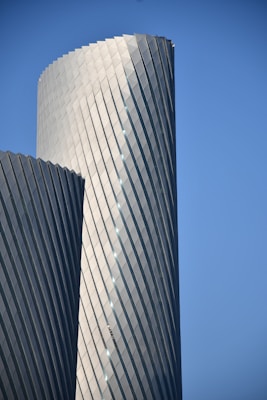 A modern skyscraper with a futuristic design is shown, featuring a twisted, spiral-like structure made up of sleek metallic panels. The building stands tall against a clear blue sky, accentuating its unique architectural style.