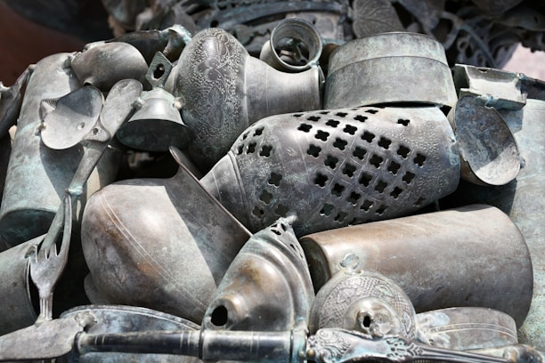 A collection of intricately designed metal objects, including vases, bowls, and a bell, arranged in a decorative manner. The items display ornate patterns and various levels of tarnish, giving them an antique appearance.