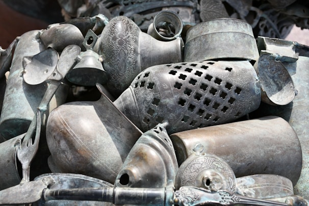 A collection of intricately designed metal objects, including vases, bowls, and a bell, arranged in a decorative manner. The items display ornate patterns and various levels of tarnish, giving them an antique appearance.