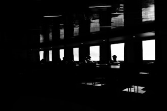 A minimalist black and white interior setting featuring silhouetted figures sitting by large rectangular windows. The high contrast lighting creates stark shadows and reflections, adding a sense of mystery and contemplation.
