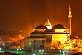 An illuminated mosque at night with a prominent minaret and several domes. The building is bathed in a warm golden light, creating striking contrasts against the dark evening sky. Sparse trees and lampposts with greenish-white lights are visible in the foreground, enhancing the serene atmosphere.