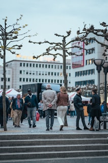 A group of people walking and interacting in an urban setting surrounded by buildings, with leafless trees lining the pathway. Some individuals appear to be engaged in conversation while others are carrying shopping bags. Tents and a truck can be seen in the background.