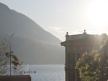 A serene lakeside view with a hotel building silhouetted against a bright sun. The hotel is positioned to the right, featuring classic architectural details. In the background, mountains rise above the calm water of the lake. The sunlight creates a hazy yet warm environment, casting a soft glow over the scene. Nearby greenery and a directional sign complete the tranquil setting.