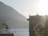 A serene lakeside view with a hotel building silhouetted against a bright sun. The hotel is positioned to the right, featuring classic architectural details. In the background, mountains rise above the calm water of the lake. The sunlight creates a hazy yet warm environment, casting a soft glow over the scene. Nearby greenery and a directional sign complete the tranquil setting.