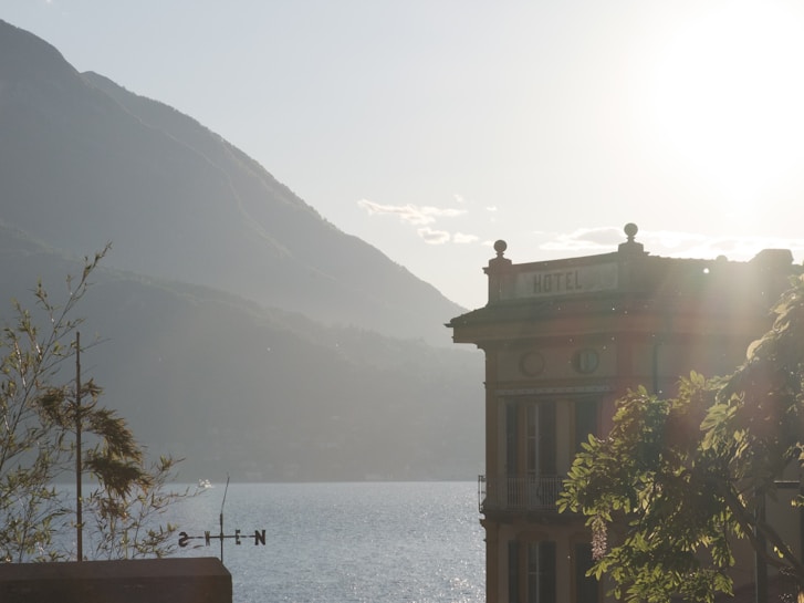 A serene lakeside view with a hotel building silhouetted against a bright sun. The hotel is positioned to the right, featuring classic architectural details. In the background, mountains rise above the calm water of the lake. The sunlight creates a hazy yet warm environment, casting a soft glow over the scene. Nearby greenery and a directional sign complete the tranquil setting.
