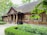 A rustic wooden house with a thatched roof, surrounded by lush greenery. There are large wooden columns at the entrance and flower pots with plants decorating the porch. The windows are symmetrically placed and have wooden shutters. Trees and bushes frame the scene, providing a serene, natural setting.
