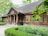 A rustic wooden house with a thatched roof, surrounded by lush greenery. There are large wooden columns at the entrance and flower pots with plants decorating the porch. The windows are symmetrically placed and have wooden shutters. Trees and bushes frame the scene, providing a serene, natural setting.