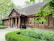 A rustic wooden house with a thatched roof, surrounded by lush greenery. There are large wooden columns at the entrance and flower pots with plants decorating the porch. The windows are symmetrically placed and have wooden shutters. Trees and bushes frame the scene, providing a serene, natural setting.