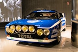 A classic rally car with prominent front spotlights and a blue and white color scheme. The car displays various brand logos and sponsors, including Lancia, Pirelli, and Total. It is showcased in a well-lit indoor environment with a black and white photograph projected on the wall behind it.