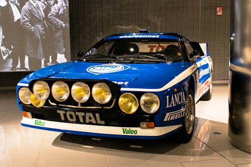 A classic rally car with prominent front spotlights and a blue and white color scheme. The car displays various brand logos and sponsors, including Lancia, Pirelli, and Total. It is showcased in a well-lit indoor environment with a black and white photograph projected on the wall behind it.