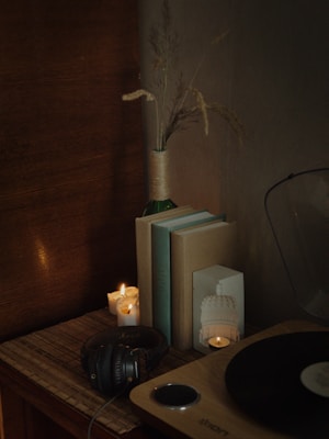 A cozy and vintage-themed setup featuring a stack of books, a pair of black headphones, a few lit candles, and a wooden turntable partially visible at the corner. A decorative vase wrapped in twine contains dried plants, adding an element of rustic charm. The ambient lighting creates a warm and inviting atmosphere.