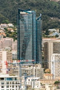 A modern high-rise building with a sleek, reflective glass facade dominates the scene. The building is surrounded by various other residential and commercial buildings that are densely clustered together. Several construction cranes are visible in the foreground, indicating ongoing construction activity. The background shows a forested hillside with scattered homes and greenery.