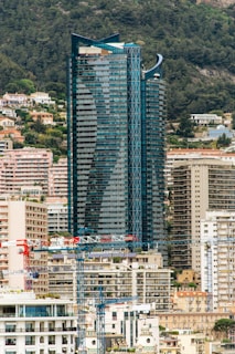 A modern high-rise building with a sleek, reflective glass facade dominates the scene. The building is surrounded by various other residential and commercial buildings that are densely clustered together. Several construction cranes are visible in the foreground, indicating ongoing construction activity. The background shows a forested hillside with scattered homes and greenery.
