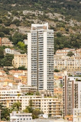 A tall, modern high-rise building stands prominently among lower residential structures and lush greenery. The background features a hillside covered with trees, enhancing the contrast between nature and urban architecture. Various smaller buildings with diverse architectural styles surround the high-rise, creating a dense cityscape.