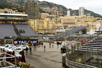Grandstands and seating arrangements are set up along a waterfront cityscape, with multiple high-rise buildings in the background. There are a few people walking and several parked cars. A TAG Heuer advertisement is prominently displayed. The scene is framed by a mountainous landscape, which adds to the urban yet scenic setting.