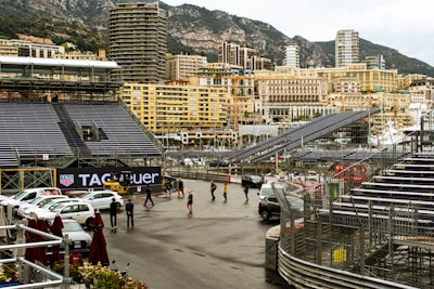 Grandstands and seating arrangements are set up along a waterfront cityscape, with multiple high-rise buildings in the background. There are a few people walking and several parked cars. A TAG Heuer advertisement is prominently displayed. The scene is framed by a mountainous landscape, which adds to the urban yet scenic setting.